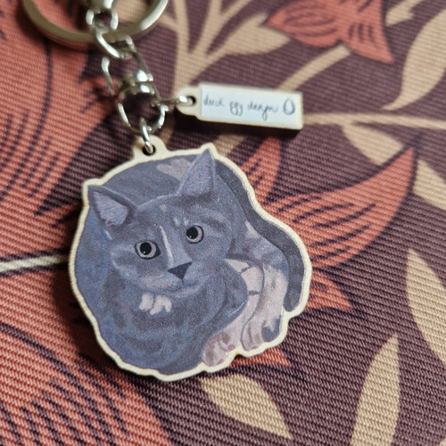 A close up of the grey and tortoiseshell cat ,earring and its small white charm with the duck egg designs logo. Behind the keyring is a retro patterned floral fabric in brown.