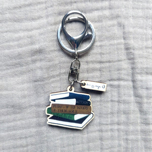 A picture of the key ring showing the silver lobster clasp as well as showing the wooden stack of book charm, and the white duck egg designs logo charm. Behind the key ring is an off grey fabric background.