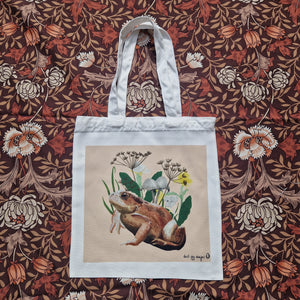 A white tote lies on a brown retro floral patterned fabric. The bag features a peach coloured square with a common toad and some wild flowers, mushrooms and grasses,. In the bottom right of the square you can see a black Duck Egg Designs logo.
