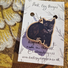 Load image into Gallery viewer, A close up view of a black and tortoiseshell cat wooden charm sits on a silver chain attached to a white backing card with a black leafy vine design. Behind the backing card you can see a warm brown retro floral fabric.
