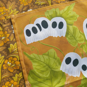 A close up of the top left of the warm orange tea towel with ghost pumpkins on a green leafy vine. Above and to the left of the tea towel you can see a warm brown retro floral patterned fabric. 