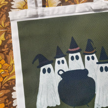 Load image into Gallery viewer, A close up of the the bag showing the coven of ghosts wearing witches hats all grouped around a cauldron in front of a green background. The bag sits on a warm yellow and borwn retro floral patterned background.

