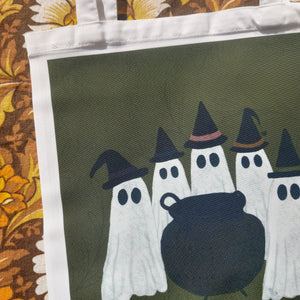A close up of the the bag showing the coven of ghosts wearing witches hats all grouped around a cauldron in front of a green background. The bag sits on a warm yellow and borwn retro floral patterned background.