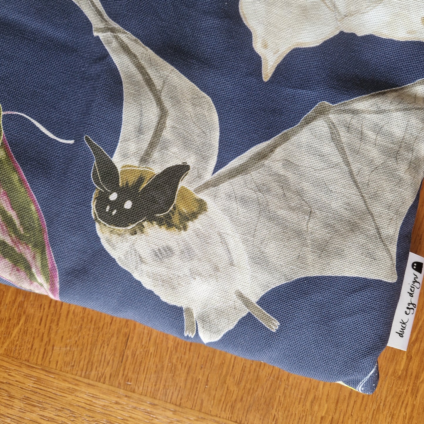 A close-up of the Duck Egg Designs Nocturnal Pollinators Small Laptop/Tablet Sleeve, made from recycled fabric with a printed cream bat on dark blue and a visible Duck Egg Designs label, shown on a wooden surface.