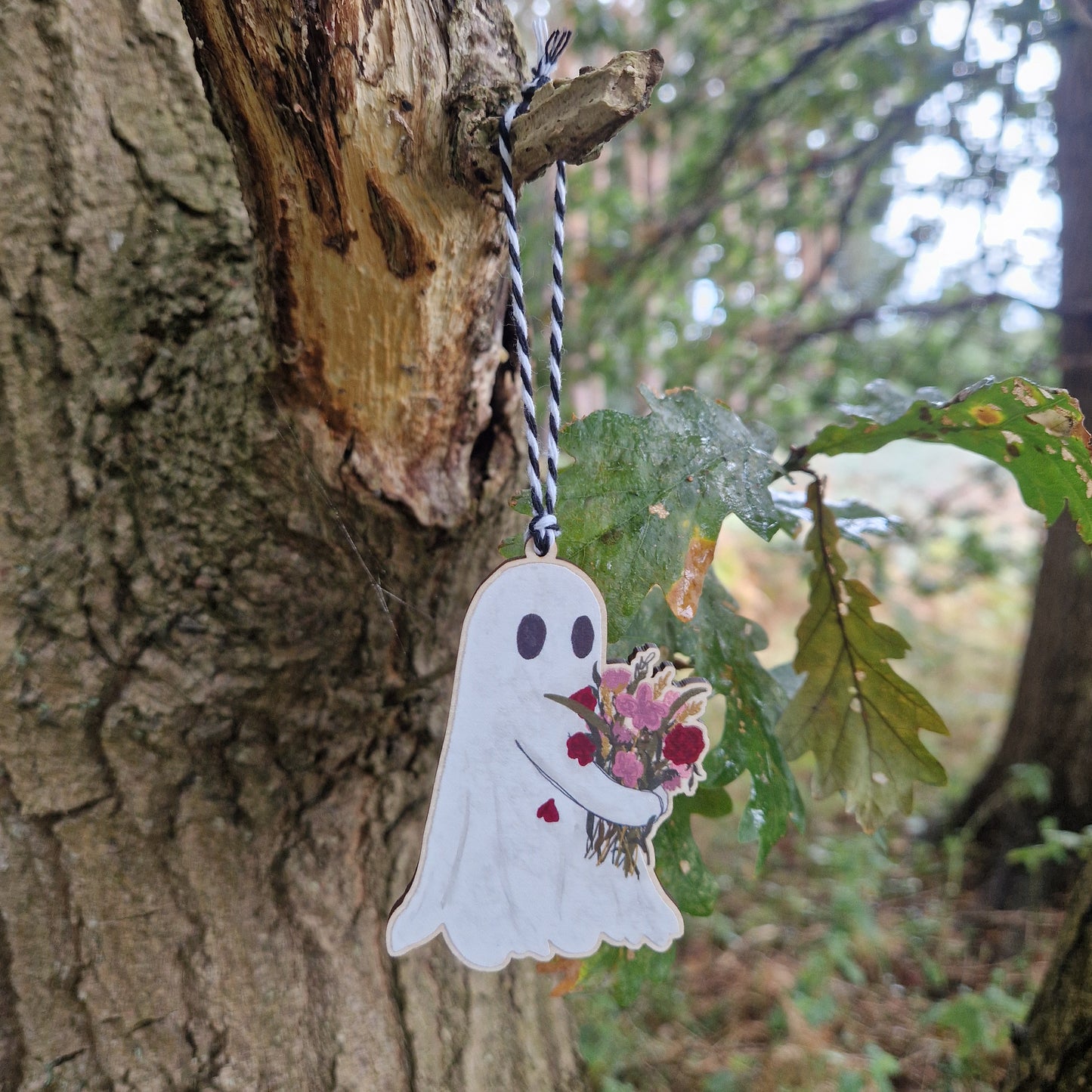 The Flower Ghost Hanging Wooden Decoration by Duck Egg Designs features an illustrated wooden ornament of a ghost holding a bouquet, hanging from a tree branch amid greenery and blurred trees in the background.