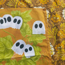Load image into Gallery viewer, The top right of the warm orange tea towel showing the ghost pumpkins surrounded by green leaves and vine tendrils. Above and to the left of the tea towel you can see a warm brown retro floral patterned fabric.
