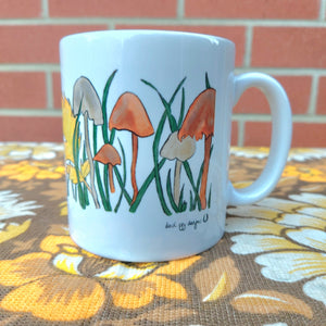 The right hand side of a white mug with a fungi filled design featuring autumn leaves and grasses too. The mug sits on a warm brown retro floral fabric in front of a red brick wall.