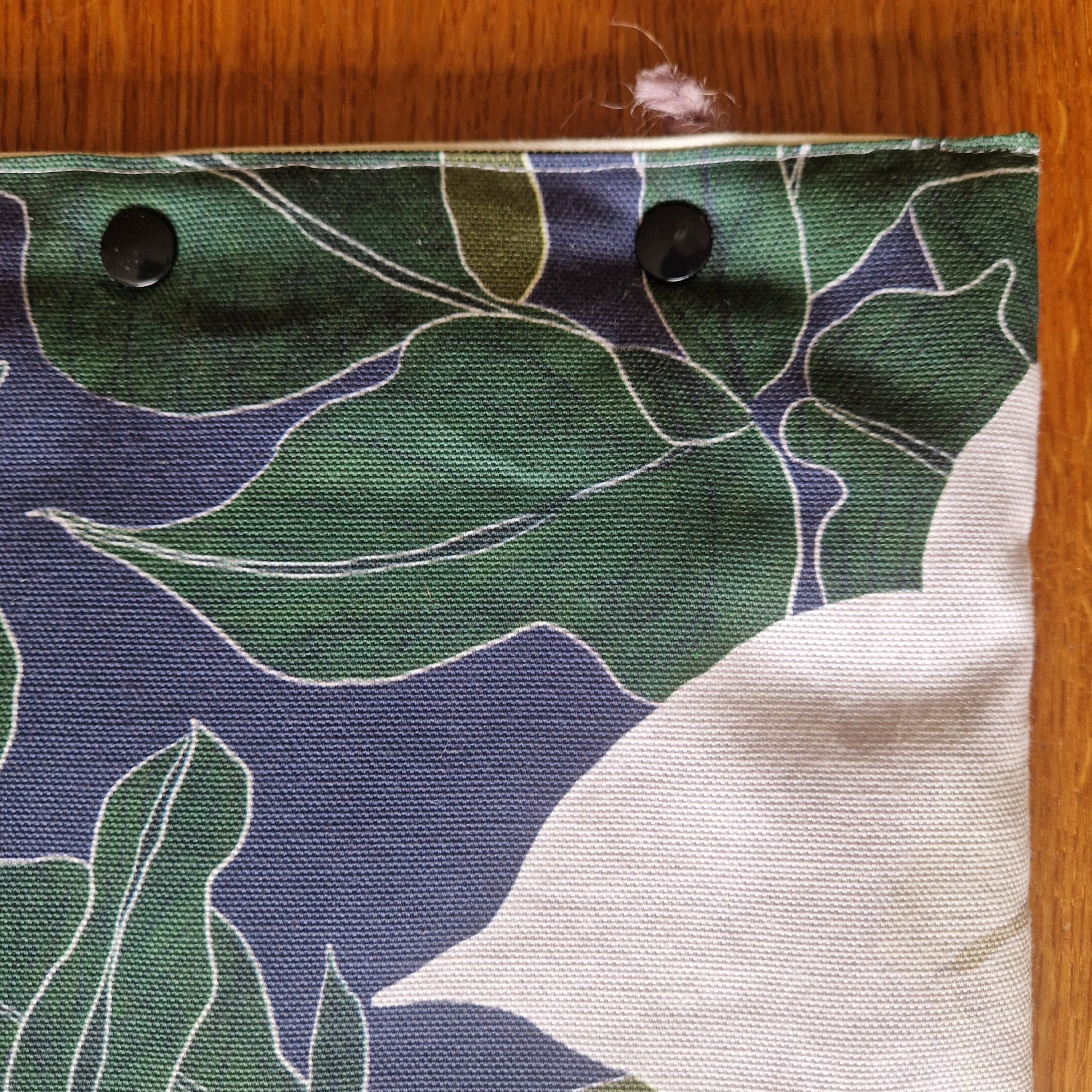 Close-up of the Nocturnal Pollinators Small Laptop/Tablet Sleeve by Duck Egg Designs, handmade from recycled fabric with a green and white leaf pattern, two black metal grommets on top, and a frayed upper right corner on a wooden background.