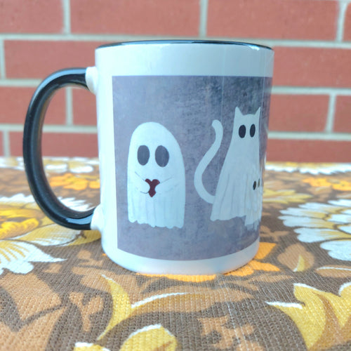 The left hand side of the mainly white mug with a black inside and handle. The mug features a ghost design on a mottled grey square background. Under the mug is a warm brown retro floral patterned fabric and it sits in front of a red brick wall.