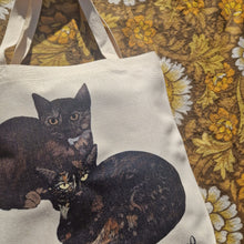 Load image into Gallery viewer, A close up view of the two black and tortoiseshell cats on the white bag showing their unique markings. To the right of the bag you can see a brown retro floral patterned fabric. 
