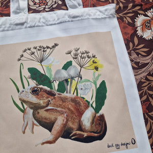 A close up view of the bag showing the detail in the toad illustration and plants surrounding him. To the right of the tote you can see a retro brown floral patterned background.