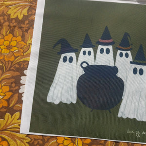 A close up of the lower left of a white bag with a green square printed onto it. The square has a group of white ghosts around a cauldron all wearing witches hats.  