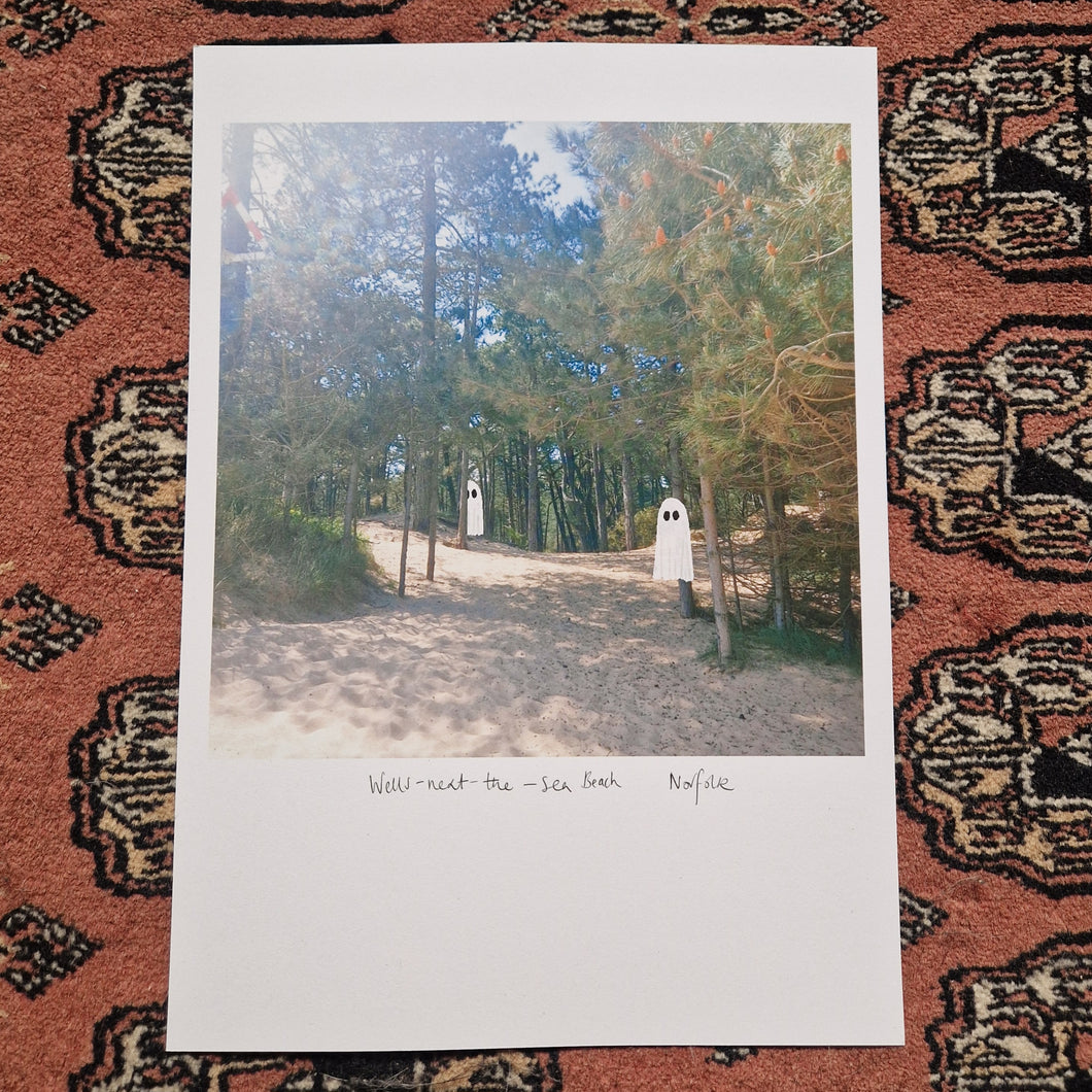 The A4 version of the ghost print sits on a red textured patterned background. The print features two ghost floating in the shade of pine trees above a sandy beach.
