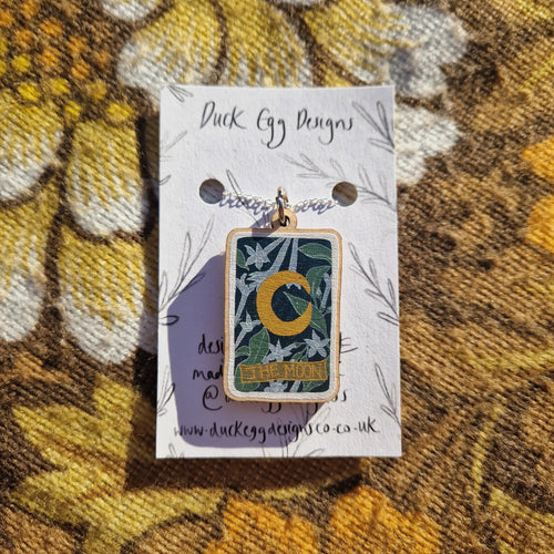 A white backing card sits on a retro brown floral patterned fabric.  The backing card has a silver necklace with a colourful wooden moon tarot design.