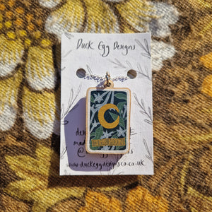 A white backing card sits on a retro brown floral patterned fabric.  The backing card has a silver necklace with a colourful wooden moon tarot design.