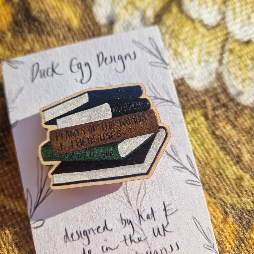 A close up view of the wooden stack of books pin badge featuring 5 different coloured books. The pin badge sits on a white backing card with the words ‘Duck Egg Designs’ in black. Behind the backing card you can see a brown retro floral patterned fabric.
