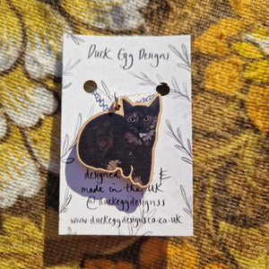 In the middle of the image is a white backing card with a black leafy vine design sitting on a warm brown retro floral fabric background. The backing card has a silver chain with a wooden curled up black and tortoiseshell cat charm.
