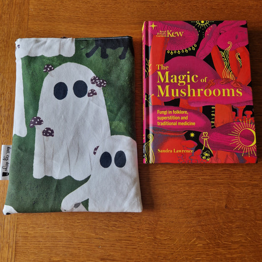 A Haunting Of Ghosts Book Sleeve by Duck Egg Designs, crafted from recycled fabric with cute ghost and mushroom illustrations, sits beside The Magic of Mushrooms by Sandra Lawrence on a wooden surface.