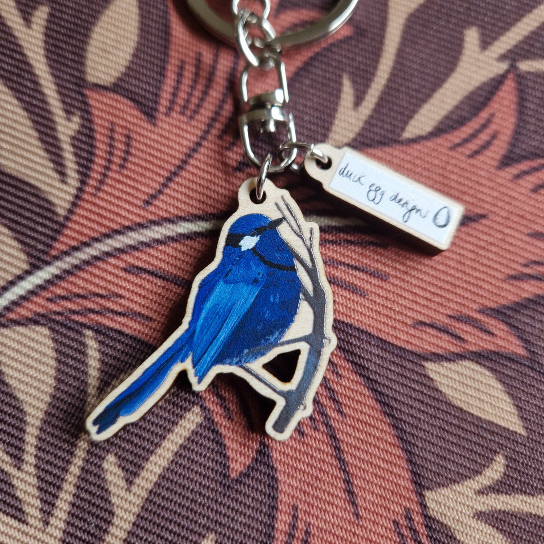 Blue fairy wren keyring with a white duck egg designs charm sits on a brown retro floral fabric.