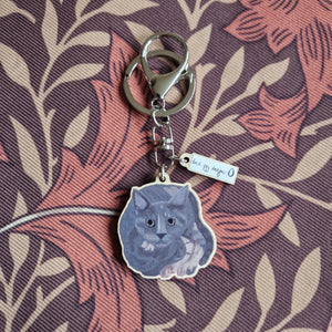 A grey and tortoiseshell cat keyring sits on a retro brown floral patterned background. He keyring has silver hardware and a small charm with the duck egg designs logo.