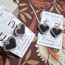 Load image into Gallery viewer, Two pairs of earrings with black heart charms on them sit at angles to each other. One pair has shepherds crook findings while the other has silver hoops. The earrings sit on white backing cards with black writing that reads ‘Duck Egg Designs’ as well as a simple leafy vine design.  Behind the backing cards you can see a brown retro floral patterned background.
