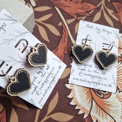 Two pairs of earrings with black heart charms on them sit at angles to each other. One pair has shepherds crook findings while the other has silver hoops. The earrings sit on white backing cards with black writing that reads ‘Duck Egg Designs’ as well as a simple leafy vine design.  Behind the backing cards you can see a brown retro floral patterned background.