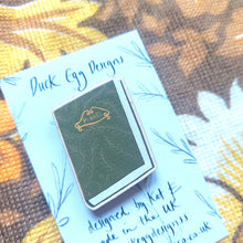 Load image into Gallery viewer, A green pin badge of a book with the word ‘PLANTS’ on the front in yellow sits on a white backing card on some floral retro patterned fabric.

