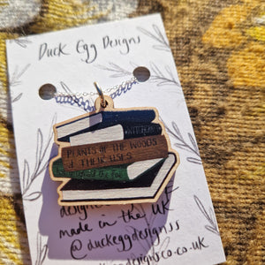 A stack of books necklace with a silver chain sits on a white backing card with a black leafy vine design. Behind the backing card is a brown floral patterned fabric. 