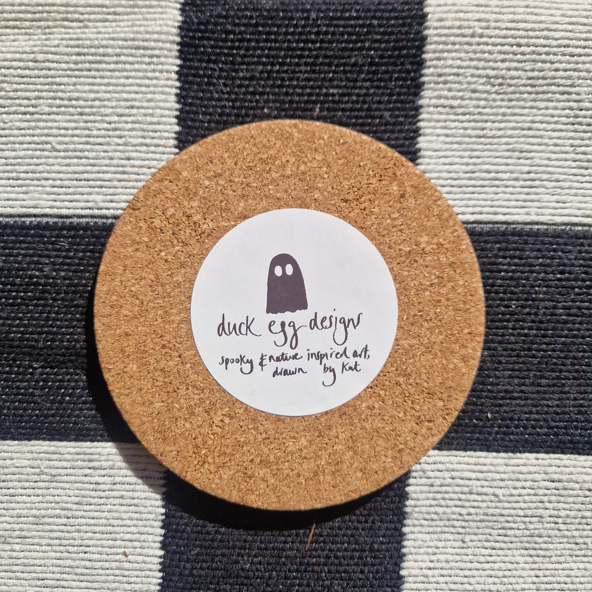The Baking Ghost coaster by Duck Egg Designs features a simple ghost illustration and text “spooky & sassy inspired art, drawn by Kat” on a round cork base. Printed in the UK and shown on black-and-white checkered fabric.