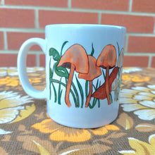 Load image into Gallery viewer, The left side of a white mug featuring a fungi filled design with long grasses and autumn leaves. The mug sits on a warm brown retro floral fabric and in front of a red brick wall.
