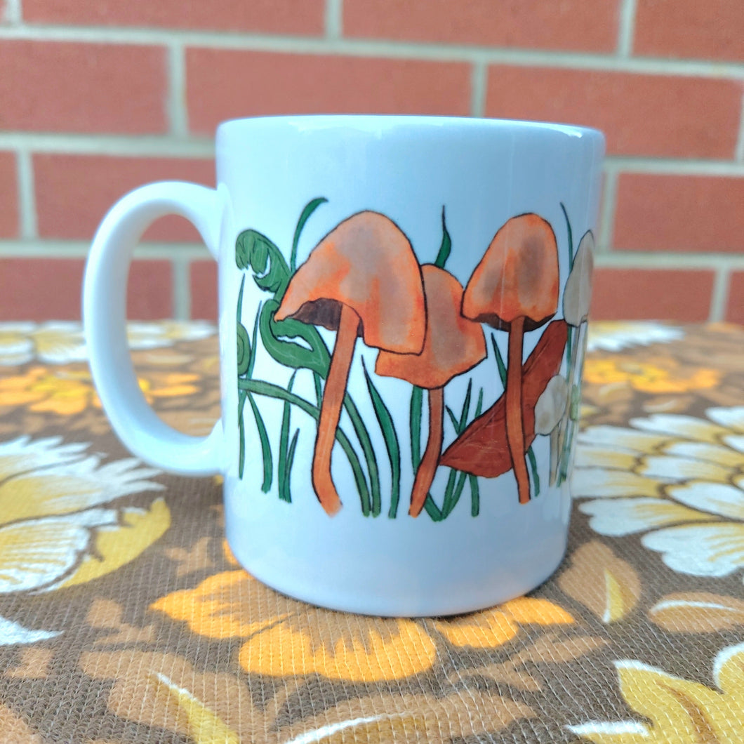 The left side of a white mug featuring a fungi filled design with long grasses and autumn leaves. The mug sits on a warm brown retro floral fabric and in front of a red brick wall.
