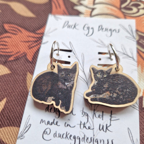 A close up of a mixed pair of black tortoiseshell cat sister earrings. The charms are on silver hoops hanging from a white backing card with a black leafy vine design and the words ‘Duck Egg Designs’ across the top. Behind the backing card you can see a retro floral patterned fabric in browns and oranges.