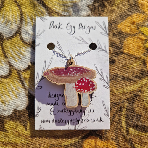 A white backing card featuring the words ‘Duck Egg Designs’ in black handwriting with a silver necklace featuring a wooden charm with two fly agaric mushrooms. Behind the backing card you can see a brown retro floral patterned background. 