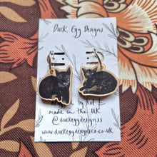 Load image into Gallery viewer, A pair of mixed cat earrings of black tortoiseshell kittens with silver hoop findings sit attached to a white backing card in front of a retro floral patterned background. The backing card features the words ‘Duck Egg Designs’ across the top and a leafy vine design.
