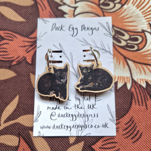A pair of mixed cat earrings of black tortoiseshell kittens with silver hoop findings sit attached to a white backing card in front of a retro floral patterned background. The backing card features the words ‘Duck Egg Designs’ across the top and a leafy vine design.