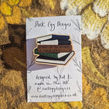 Load image into Gallery viewer, A white backing card with black writing on sits on a retro floral brown patterned fabric. The backing card has a wooden pin badge with a stack of books design.
