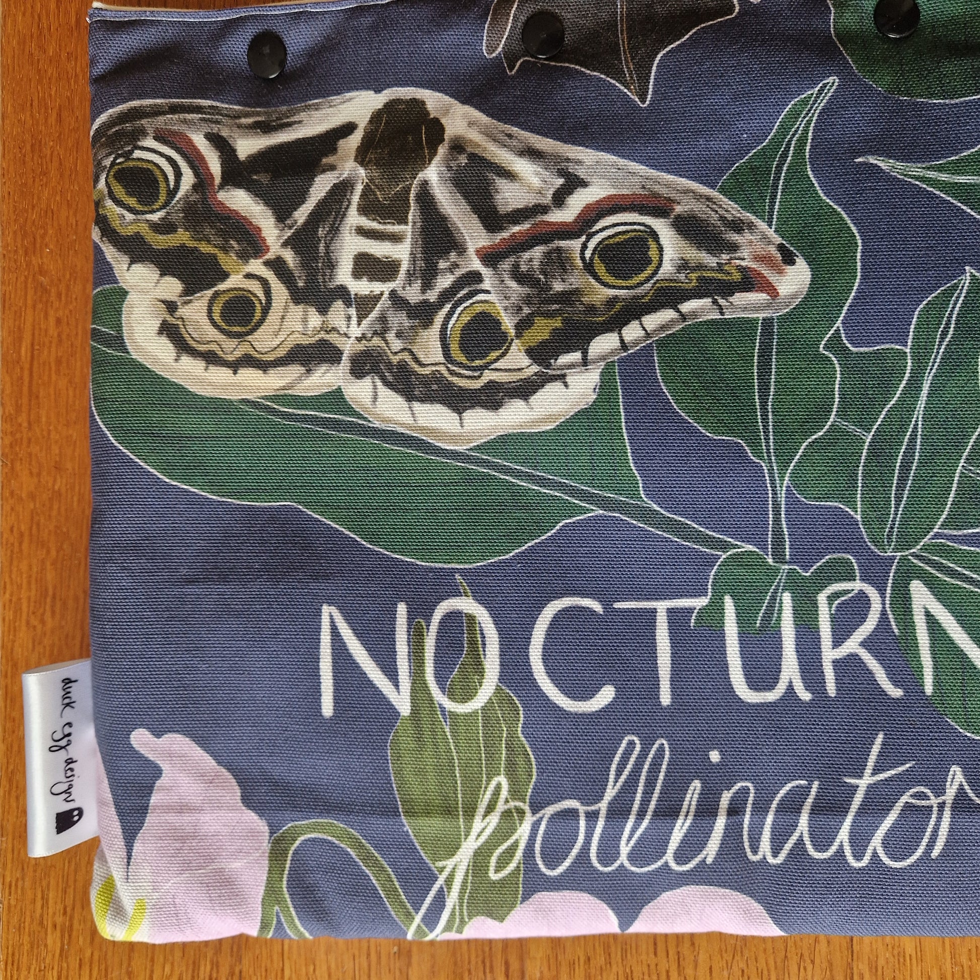 The Duck Egg Designs Nocturnal Pollinators Small Laptop/Tablet Sleeve is handmade from recycled fabric, featuring a moth, green leaves, the words NOCTURN… pollinato…, and a small white side tag. It rests on a wooden surface.