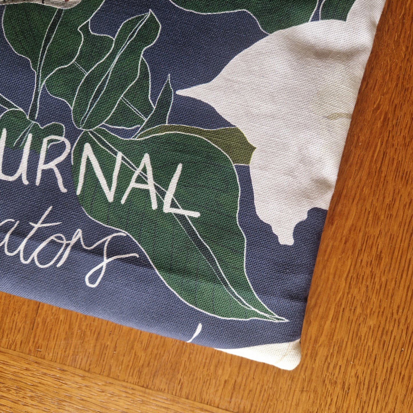 A corner of the Nocturnal Pollinators Small Laptop/Tablet Sleeve by Duck Egg Designs, featuring green leaves, white flowers, and partial text on a dark blue background, rests on a wooden surface.