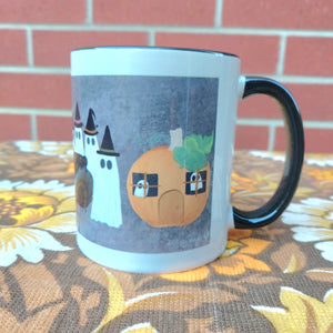 The right side of the mug is on display showing off the black handle and rim as well as the ghosts on that side of the mug. The mug sits on a warm brown floral patterned fabric, and in front of a red brick wall. 
