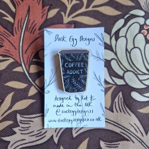 A black coffee cup wooden pin badge with the words ‘Coffee Addict’ in white handwriting and a leafy vine design sits on a white and black backing card. Behind the backing card you can see a retro brown floral patterned fabric.