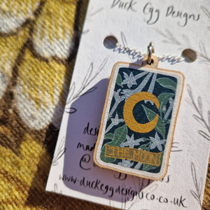 A close up of a wooden moon tarot charm with a white, blue, green and yellow design. The charm hangs from a sterling silver chain which is attached to a white backing card which lies on a brown retro floral patterned fabric.