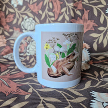 Load image into Gallery viewer, The view of the side of a mug featuring a toad with fungi and wildflowers behind them on a pale peachy coloured background. Behind the mug is a retro floral patterned fabric.
