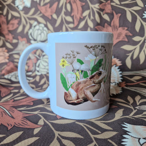 The view of the side of a mug featuring a toad with fungi and wildflowers behind them on a pale peachy coloured background. Behind the mug is a retro floral patterned fabric.