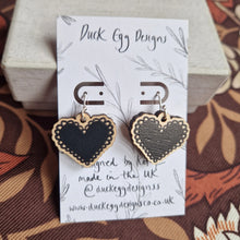 Load image into Gallery viewer, A white backing card with a pair of black heart earrings with shepherds crook findings leans against a cream coloured box. Under the box and behind the backing card is a retro floral patterned fabric.
