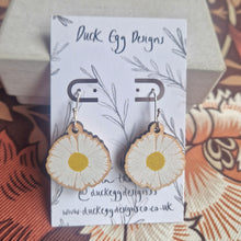 Load image into Gallery viewer, A pair of daisy earrings with silver shepherds crook findings sit on a white and black backing card leant against a cream box. The backing card features the words ‘Duck Egg Designs’ in black handwriting as well as a leafy vine pattern. In the background you can see a retro floral patterned fabric in browns and oranges.
