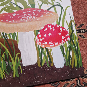 A close up of the bottom right of the print showcasing the detail in the artwork. You can see two of the fly agaric toadstools as well as green grasses and the brown soil.  To the right and bottom of the print you can see a red textured patterned background. 