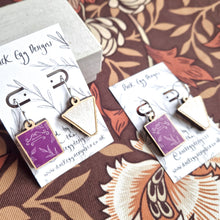 Load image into Gallery viewer, Two pairs of earrings sit on white and black backing cards next to each other. Both pairs feature a mismatched pair of books - a purple book on the left with a leafy design and the word BOTANICAL in white, and an open book sat on its spine on the right. The left hand pair has shepherds crook finding, while the right pair has silver hoops. Behind the earrings is a brown retro floral patterned background. 
