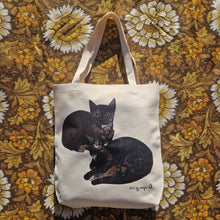 Load image into Gallery viewer, A white textured tote bag sits on a brown floral retro patterned background. The bag features two black and tortoiseshell cats curled up next to each other, with a black duck egg designs logo in the bottom right of the bag.
