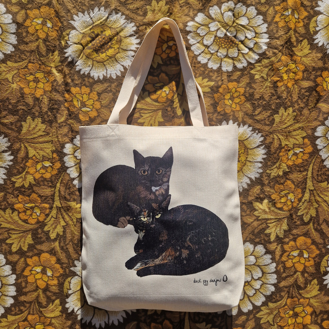 A white textured tote bag sits on a brown floral retro patterned background. The bag features two black and tortoiseshell cats curled up next to each other, with a black duck egg designs logo in the bottom right of the bag.