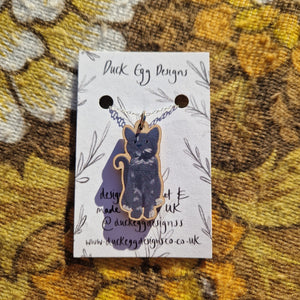 In the middle of the image is a white backing card featuring a black leafy vine design. Hanging from a silver chain attached to the backing card is a grey cat charm featuring a sitting kitten. Behind the backing card you can see a warm brown retro floral patterned fabric.